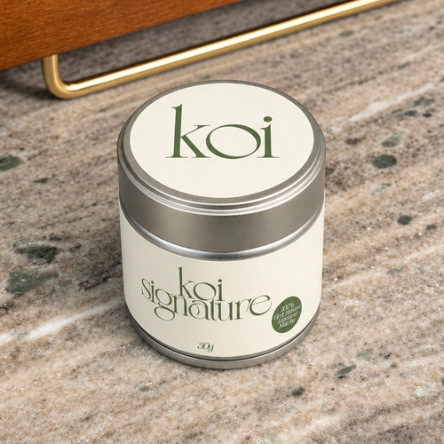 A small round tin candle labeled 'koi signature' sits on a speckled stone countertop with a wooden shelf and brass handle in the background.